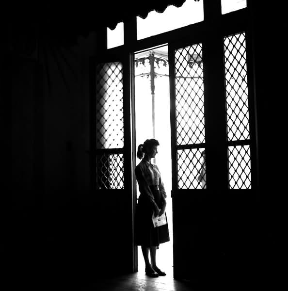 Foto de uma porta com uma mulher ao centro. A foto é escura com um claro bem no meio, onde a porta está aberta.