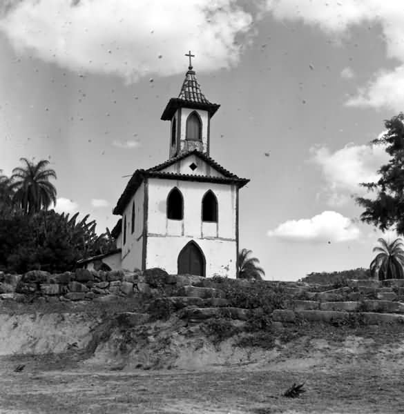 Foto em preto e branco com igreja estilo colonial em destaque, ao fundo da igreja bananeiras e palmeiras, a frente uma escada de pedras.
