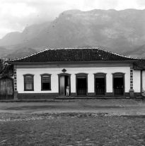Uma casa colonial em Catas Altas, Minas Gerais é mostrada de frente, com suas quatro portas e duas janelas. Um homem está de pé encostado em uma das portas. Por trás da casa avista-se a serra, parcialmente oculta pela neblina. A foto foi tirada em 1960 pelo fotógrafo Mazonnni.
