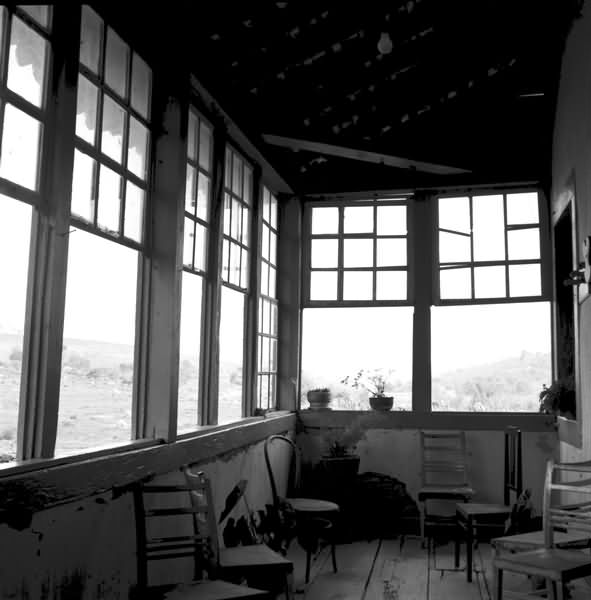 Imagem em preto e branco da varanda de uma fazenda, coberta em forma de L, com sete janelas retas, abertas, com vidros quadriculados. Tem sete cadeiras espalhadas pela varanda, vasinhos de plantas em uma das janelas e em alguns cantos do ambiente.
