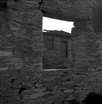Vista da janela de uma residencia feita em pedra, que mostra uma outra residencia antiga ao fundo construída também com pedras sobrepostas com janelas e portas de madeira.