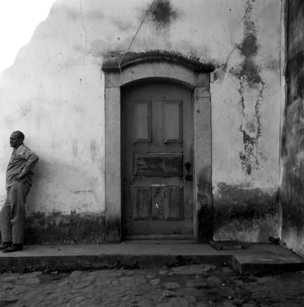 Construção em ruína, com uma porta fechada e homem encostado, com mãos na cintura, na parede com um olhar direcionado ao horizonte.