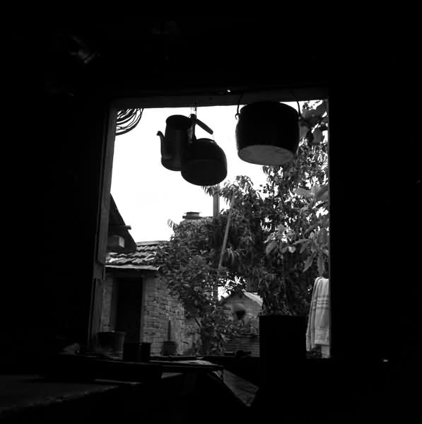 Imagem em preto e branco de uma janela. Há uma chaleira e duas panelas penduradas na parte superior da janela. Ao fundo há uma outra casa de tijolos que está com a porta aberta, do lado direito da imagem tem a folhagem de uma árvore e um varal em que possui uma toalha pendurada.